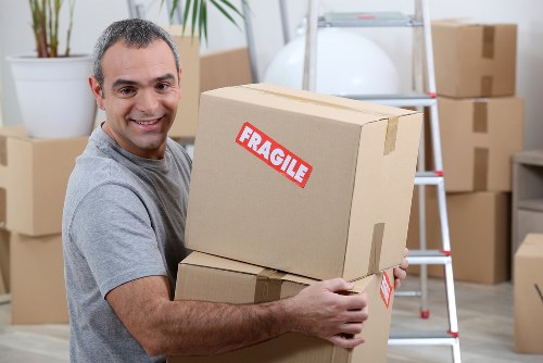 A smiling man with short dark hair is holding two cardboard boxes, with one stacked on top of the other, in a room that appears to be part of a residential property. The boxes are sealed with packing tape, and one features a red 'FRAGILE' label, indicating they contain delicate items. In the background, there are several other cardboard boxes, some stacked on the floor and others on a step ladder or leaning against the wall, suggesting an ongoing packing or moving process. The room has light-colored walls and a white ladder, and a tall potted plant is visible on the left side, adding a touch of greenery. The scene is well-lit with natural or artificial light, creating a clear and professional atmosphere typical of house removals services in the Waterloo or surrounding areas. The overall setting reflects a typical interior during a move, with household items ready for transportation, consistent with the services offered by Waterloo Man with Van in the local area near [POSTCODE].