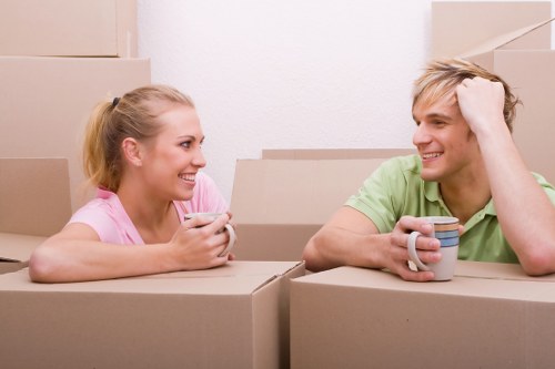 A young woman and a young man are seated inside a residential property surrounded by stacked cardboard moving boxes, indicating a house move in progress. The woman, with blonde hair tied back and wearing a pink top, is smiling and holding a smartphone, while the man, with blonde hair and dressed in a light green shirt, is also smiling, holding a white ceramic mug in his right hand and touching his hair with his left hand. The space appears to be a living room or hallway, with plain white walls and natural lighting. The cardboard boxes vary in size, some sealed with packing tape, and are arranged in the foreground and background, suggesting an active packing or unpacking process. The environment is tidy, and the scene captures a moment of casual conversation and preparation amidst the household removals, consistent with services offered by Waterloo Man with Van in the Waterloo area near London.