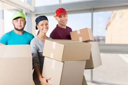 The image depicts three professional movers from Waterloo Man with Van working indoors in a well-lit area, possibly a house or flat in Waterloo, London. The movers are dressed in casual work attire, each wearing a different brightly coloured cap—one in green, one in navy, and one in red—conveying a friendly and approachable service. The male mover in the red cap is smiling and holding a medium-sized brown cardboard box, while the female mover in the navy cap is also smiling and carrying a similar box. Behind them, a fourth individual, a male with a beard, is partially visible, wearing a green cap and holding a large cardboard box. The scene is set against a backdrop of large windows with natural daylight illuminating the workspace, revealing a residential street outside. The focus is on the safe handling and transportation of household items, including various cardboard boxes of different sizes. The environment suggests a professional moving service in the Waterloo area, specializing in residential relocations, with an emphasis on efficiency and customer readiness.