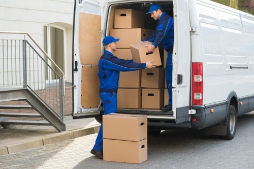 Two professional movers from Waterloo Man with Van, dressed in blue uniforms and caps, are loading cardboard boxes into the rear of a white removals van parked outside a residential building. One mover is inside the van, passing a box to the other who is standing outside on the pavement. The boxes are standard brown cardboard, stacked neatly both inside and outside the van, with some on the ground awaiting transfer. The setting appears to be a suburban street in the UK, with the edge of a building and a staircase visible to the left, indicating the house has a small entrance porch accessible via external stairs. The interior of the van is open, showing more boxes arranged for transportation. The lighting suggests daytime, with natural light illuminating the scene. The action reflects typical house removals, highlighting careful handling of household belongings in a professional manner, aligned with services offered by Waterloo Man with Van for local relocations within the London area or nearby districts.