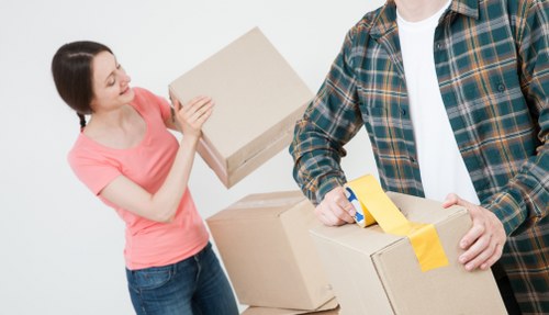 A woman with dark hair pulled back, wearing a pink T-shirt and jeans, is smiling and holding a plain cardboard box, appearing to be preparing for a house move. Beside her, a man in a plaid shirt over a white T-shirt is holding another large cardboard box with yellow packing tape around it, suggesting they are in the process of packing or unloading belongings. Several other cardboard boxes of various sizes are stacked around them, some open, revealing items inside. They are positioned against a plain white background, indicating an indoor setting, possibly a residential interior. The scene is lit evenly, highlighting the textures of the cardboard, fabric, and the individuals' clothing, consistent with professional house removals activities carried out by Waterloo Man with Van in the local area. The image conveys efficiency and cooperation typical of local house removal services in the Waterloo or nearby postcode area.