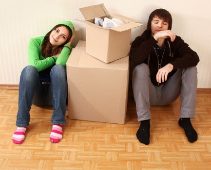 A young woman with long, brown hair, wearing a bright green hoodie and blue jeans, sits on a wooden floor with her head resting on her arm, appearing tired or bored. Next to her, a young man with dark brown hair, dressed in a black hoodie and grey jeans, sits with his arms resting on his knees, looking down pensively. Between them is a stack of moving boxes, including a large cardboard box on the floor and an open, smaller box placed on top, containing packing paper. The setting appears to be an indoor residential space, with a plain light-colored wall behind them and a smooth wooden floor, typical of a house or apartment in the Waterloo area of London. This scene reflects the process of packing during a house move, relevant to Waterloo Man with Van's removals services in London postcodes beginning with SW or SE.