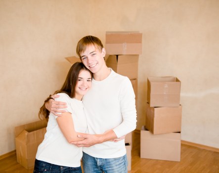 A young couple with light skin and casual clothing, standing inside a residential property, are smiling and embracing each other in front of a beige wall. Behind them are several cardboard moving boxes of varying sizes, some stacked on top of each other, indicating a recent or ongoing house move. The boxes are plain brown and sealed with packing tape. The man has short, light brown hair and is wearing a white long-sleeved shirt and blue jeans. The woman has long, dark brown hair and is dressed in a white short-sleeved top and dark jeans. The scene suggests a house removal or relocation context, typical of services offered by Waterloo Man with Van in the London area. The lighting is natural and even, illuminating the entire scene clearly, with the focus on the couple and the packed boxes in a tidy, carpeted or wooden-floored interior, suitable for a house moving service webpage related to the London postcode area.