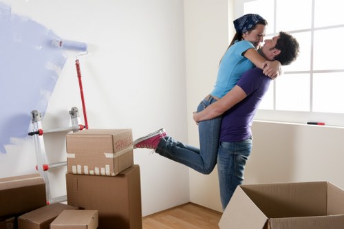 In a bright, minimally decorated room with white walls and a large window, a woman wearing a blue t-shirt, dark jeans, and red shoes is being lifted by a man wearing a purple t-shirt and dark jeans. They are smiling and embracing each other, with the woman holding onto the man's shoulders and the man supporting her around the waist. The room contains several cardboard boxes, some stacked on top of each other, indicating a moving or packing activity. A partially unrolled roll of white packing tape is mounted on the wall on a dispenser, and a red-handled tool or appliance is visible beside it. The scene suggests a house relocation, consistent with removals services offered by Waterloo Man with Van, in a residential area near Waterloomanwithvan.co.uk's service coverage area. Natural daylight fills the space through the window, creating a cheerful and functional atmosphere for the moving process.