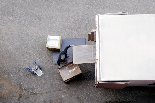 An aerial view of a person, dressed in dark clothing, loading wooden furniture into a large white removals van parked on a residential street. The furniture includes a light-colored wooden table, a smaller dark wooden side table, and a grey upholstered armchair with wooden legs. Several cardboard boxes of different sizes are nearby, some closed and one open, revealing contents. On the concrete pavement, there are also a two-stack of plastic storage containers with lids. The scene is set outdoors during daytime, with natural lighting, and the surroundings are typical of a residential area in Waterloowithvan.co.uk. The focus is on the process of loading household items, indicative of house removals or moving services, with the van positioned close to the property entrance and the items arranged for efficient packing and loading.