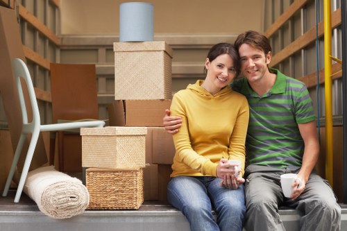 A young couple with light skin sitting together on the floor of a house removal storage area, surrounded by cardboard boxes and packing materials. The woman is wearing a yellow hoodie and jeans, holding a mug, and smiling at the camera. The man is wearing a green and purple striped polo shirt and grey trousers, also smiling and holding a mug. Behind them, there are stacked cardboard boxes of various sizes, some with textured finishes, a rolled-up carpet on the floor, a white chair leaning against the wall, and a small beige box with a blue cylinder on top. The setting appears to be a residential garage or storage space with wooden shelving units visible to the sides, indicating a house removal process in progress, possibly in the Waterloo or surrounding area. The scene is well-lit with natural or overhead lighting, emphasizing the organized packing of household items ready for transport by Waterloo Man with Van.