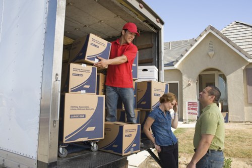 A professional removals team from Waterloo Man with Van is loading a moving van parked in front of a residential house on a suburban street. The image depicts a young man in a red cap and red shirt inside the van, lifting and stacking several large cardboard boxes labeled with the Waterloo Man with Van logo, indicating they contain household items for a house move. The boxes are mostly brown with blue printing, and some are stored on a trolley within the van's interior. Outside, a woman and a man are engaged in conversation, standing on the driveway near the open rear door of the van, with the woman wearing a dark blue shirt and the man in a light green polo shirt. In the background, the house features a light-colored facade with a pitched tiled roof and a small front garden area. The scene is well-lit with natural daylight, emphasizing the professional approach of the removals service in a typical UK residential setting, with subtle hints of Surrey postcode or local area.