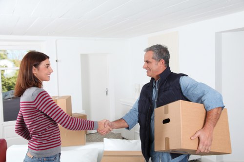 A professional man from Waterloo Man with Van, dressed in a dark blue vest and light blue shirt, is inside a bright, modern residential living room with white walls and a large window allowing natural light. He is smiling and shaking hands with a young woman wearing a red and white striped sweater, who is also smiling. The man is holding a cardboard box in his left hand, while the woman stands beside a stack of several other cardboard boxes on a white sofa or table. In the background, there are additional cardboard boxes, suggesting a house move or relocation process. The scene: a safe and organized interior space during a house removal in the Waterloo area, with a clean, minimal decor emphasizing the professional handling of household items, including the boxes and potential furniture. The lighting is natural and soft, contributing to an atmosphere of cooperation and readiness for the move, consistent with Waterloo Man with Van’s local removals service and sustainable approach.