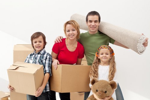 A family of four, consisting of a man, woman, son, and daughter, standing together in a bright, minimalistic interior space, likely a room within their home, with plain white walls. The man is holding a large, beige rolled-up carpet over his shoulder, while the woman is smiling and holding a medium-sized cardboard box. The boy, dressed in a checkered shirt, is holding two smaller cardboard boxes, and the girl, wearing a colourful headband and a white sweater, is holding a brown teddy bear. Several other cardboard boxes of various sizes are arranged around them, indicating a packing or moving process. The lighting is natural and evenly distributed, enhancing the scene's clarity. This setting reflects a typical interior environment where house removals or relocation activities are taking place, aligning with the services offered by Waterloo Man with Van in the context of local removals in the [POSTCODE] area, possibly near [TOWN]. The family appears organized and prepared for a move, emphasizing a professional and family-friendly approach to home relocations.