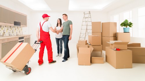 In a bright, spacious room with large windows allowing natural light, two individuals are engaged in a handshake, likely sealing a deal related to house removals. One person is dressed in red overalls and a cap, indicative of a professional mover from Waterloo Man with Van, while the other two are casually dressed in jeans and t-shirts, standing beside a ladder. The room contains a variety of packed cardboard moving boxes of different sizes, some stacked on the floor and others arranged along the walls, ready for relocation. On the left, a flat-fronted kitchen area with light wooden cabinetry and modern appliances is visible. A small potted plant sits on top of one stack of boxes near the window, adding a touch of greenery. The space appears orderly and prepared for a house move, demonstrating a typical interior scene of a professional removals service in a residential setting, with a focus on efficient packing and logistics in the area near Waterloo, London, or nearby postcode regions.