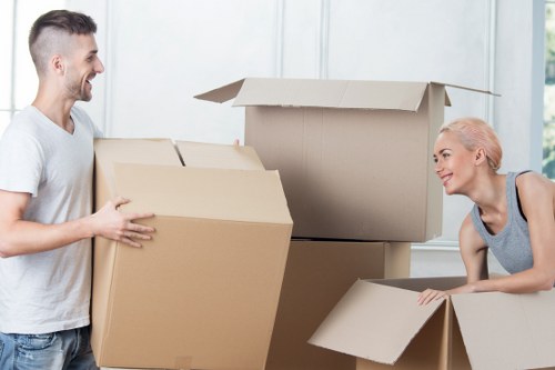 A young man and a woman with light skin and blonde hair are engaged in a house removal inside a bright, modern home with white walls and large windows. The man, dressed in a light grey T-shirt, is smiling and holding a medium-sized cardboard box, while the woman, wearing a grey sleeveless top, is leaning over a smaller cardboard box, appearing ready to pack or move items. Behind them, there are several other open cardboard boxes of various sizes, some empty and some partially filled, stacked on the floor. The boxes, made of plain brown kraft material, are arranged in a way that suggests a packing or moving process. The natural daylight streaming through the windows creates a well-lit environment, emphasizing the clean and organized space typical of residential interiors in the Waterloo or London area. This scene represents a typical house removal activity, with the focus on careful handling and packing of household belongings during a relocation, with Waterloo Man with Van providing professional removals services in the local area.