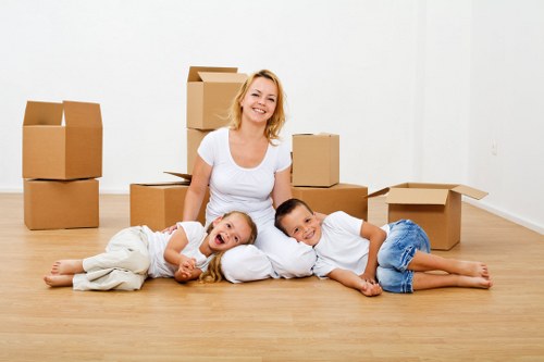 A smiling woman dressed in a white t-shirt is sitting on a light wooden floor in a bright, minimally furnished room, surrounded by several brown cardboard boxes of various sizes, some open and others closed. She is accompanied by two children lying beside her; a girl with long blonde hair and light clothing is lying on her side, resting her head on her hand, while a boy in a white t-shirt and blue jeans is lying on his stomach nearby. The background features a plain white wall, with some boxes stacked in the corner, suggesting a house move or packing process. The scene is well-lit with natural light, creating a warm and inviting atmosphere, and subtly hints at residential interior spaces typical of a house in Waterloo or the surrounding area, aligning with professional house removals services provided by Waterloo Man with Van.