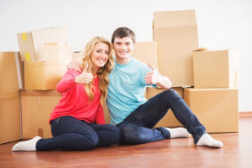 A young couple, a woman with long blonde hair wearing a red sweater and dark jeans, and a man with short dark hair wearing a light blue t-shirt and dark jeans, are seated on a wooden floor inside a residential property. They are smiling and giving thumbs-up signs, indicating satisfaction with their move. Behind them are several stacked cardboard moving boxes of varying sizes, some sealed with packing tape, arranged against a plain white wall, suggesting a house or flat in an urban area in or near Waterloo. The background shows a clean, uncluttered environment typical of a recently packed or partially unpacked room, with soft, natural lighting illuminating the scene. The couple appears relaxed and happy, symbolising a successful house removal or relocation carried out by Waterloo Man with Van, a professional removals service in the local area. This setting emphasizes the process of packing and moving household belongings, including boxes for personal items, within a residential context in South West London, near Waterloo.