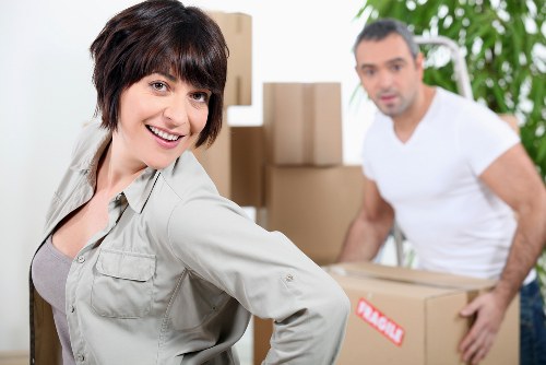 A woman with short dark hair, smiling and looking directly at the camera, is in the foreground of this image taken during a house move in a residential property. She is wearing a light-colored casual jacket over a grey top. Behind her, a man with short dark hair and a beard, dressed in a white t-shirt, is slightly out of focus and appears to be in the process of lifting or carrying a cardboard box labeled 'fragile.' The background features a house interior with beige walls and a white door frame, along with several stacked cardboard boxes of varying sizes, some sealed with packing tape, and likely containing household items ready for relocation. To the right, a houseplant with broad green leaves is visible, enhancing the domestic atmosphere. The lighting is bright and natural, suggesting daytime, and the scene captures a typical moment of residential house removals, reflecting the professional services of Waterloo Man with Van in the context of local removals across the London area.