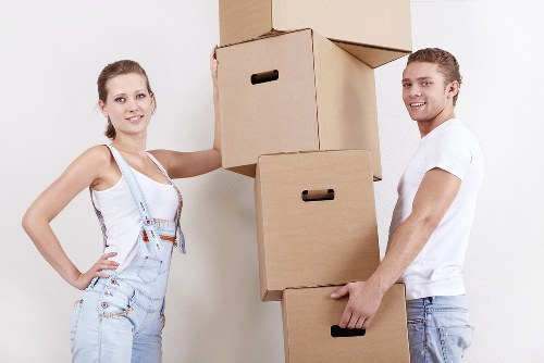A young woman and a young man are standing indoors against a plain white wall, engaged in a house move. The woman, positioned on the left, has light brown hair tied back and is wearing a white tank top and light denim overalls with a measuring tape draped around her neck. She is smiling at the camera with her left hand resting on a stack of three large cardboard boxes, which are placed one on top of the other, with the top box slightly tilted. The man, standing on the right, has short light brown hair and is wearing a white t-shirt and light blue jeans. He is also smiling at the camera and appears to be in the process of lifting or carrying the bottom box with his right hand, while his left hand is holding onto the middle box. The background suggests an interior residential environment, relevant to house removals, with smooth, neutral-colored walls. The scene subtly evokes a house clearance or relocation effort, consistent with professional removals services provided by Waterloo Man with Van for local moves within the [POSTCODE] area near [TOWN]. The natural lighting highlights the textures of the cardboard boxes and casual clothing, emphasizing the practical, systematic approach involved in moving household items efficiently and securely.
