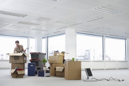 A woman in casual clothing is seen inside a modern, well-lit office space with large windows, surrounded by various cardboard boxes of different sizes, some sealed with packing tape and others open, revealing contents such as papers, books, and small household items. The boxes are stacked and scattered across the floor, with some positioned on top of each other, indicating an active packing or unpacking process during a house or office move. There is a black office chair on the carpeted floor near the boxes, along with a tangled power cable. The background reveals a bright, spacious room with white walls and a suspended ceiling equipped with fluorescent lighting. The office environment appears tidy but temporarily disorganized, reflecting a typical relocation scene coordinated by house removals services like Waterloo Man with Van, aiming to facilitate smooth household or office transitions in the local Waterloo or South London area, near postcode SE.