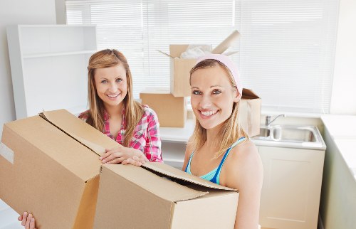 Two smiling women are engaged in packing and moving boxes within a bright, modern kitchen. One woman on the left, wearing a pink checked shirt, is holding a large cardboard box, while the other woman on the right, dressed in a blue top with a pink headband, is also handling a cardboard box, possibly preparing it for transport. The kitchen background features a stainless steel sink, a white cupboard, and a row of large windows with natural light streaming through, illuminating the space. Several cardboard boxes are stacked or placed on the floor, indicating a packing or moving process, with some open boxes showing the contents. The overall environment suggests an indoor residential setting in a house or flat in Waterloo, with a focus on domestic packing activities related to relocations or removals. Waterloo Man with Van's professional removals service is reflected in the scene through the organized setup and the emphasis on household packing tasks.