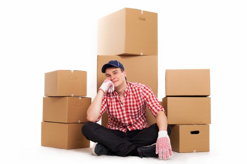 A young man dressed in a red and white checkered shirt, black trousers, and a navy blue cap sits on the floor indoors, surrounded by several brown cardboard boxes of varying sizes, some stacked behind him and others placed on either side. He is holding a pink striped cloth glove on his right hand and resting his head on his right hand, with a relaxed expression. The boxes appear to be packed for a house move, with handles visible on some. The scene is well-lit with a plain white background, indicating a controlled environment typical of professional removals or packing services. This setting suggests a house relocation scenario, consistent with services offered by Waterloo Man with Van, operating in the Waterloo area, within the London Borough of Lambeth, hinting at the South West London locale. The arrangement emphasizes the household packing aspect of a professional removals service, with focus on the boxes and the individual preparing for a move.