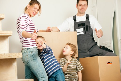 A family preparing for a house move in a well-lit, modern interior space, with a woman wearing a striped shirt and jeans, and a man dressed in work overalls, standing near a large cardboard box. The woman is giving a thumbs-up and smiling at the camera, while the man is also smiling and giving a thumbs-up gesture. Two young children, a boy in a blue and white striped shirt and a girl in a brown and white striped shirt, are positioned in front of the woman, with the boy making a playful gesture towards the camera and the girl looking up towards the woman. Visible is a partially stacked pile of cardboard boxes of various sizes, some of which are open, and the background includes an indoor staircase with wooden steps, suggesting a typical residential property in the Waterloo or broader London area. The scene captures a moment of packing and moving, reflecting the services offered by Waterloo Man with Van within the removals sector, with the overall scene emphasizing family, home contents, and professional moving efforts, set in a clean, organized environment suggesting careful handling and safety during the house removal process.